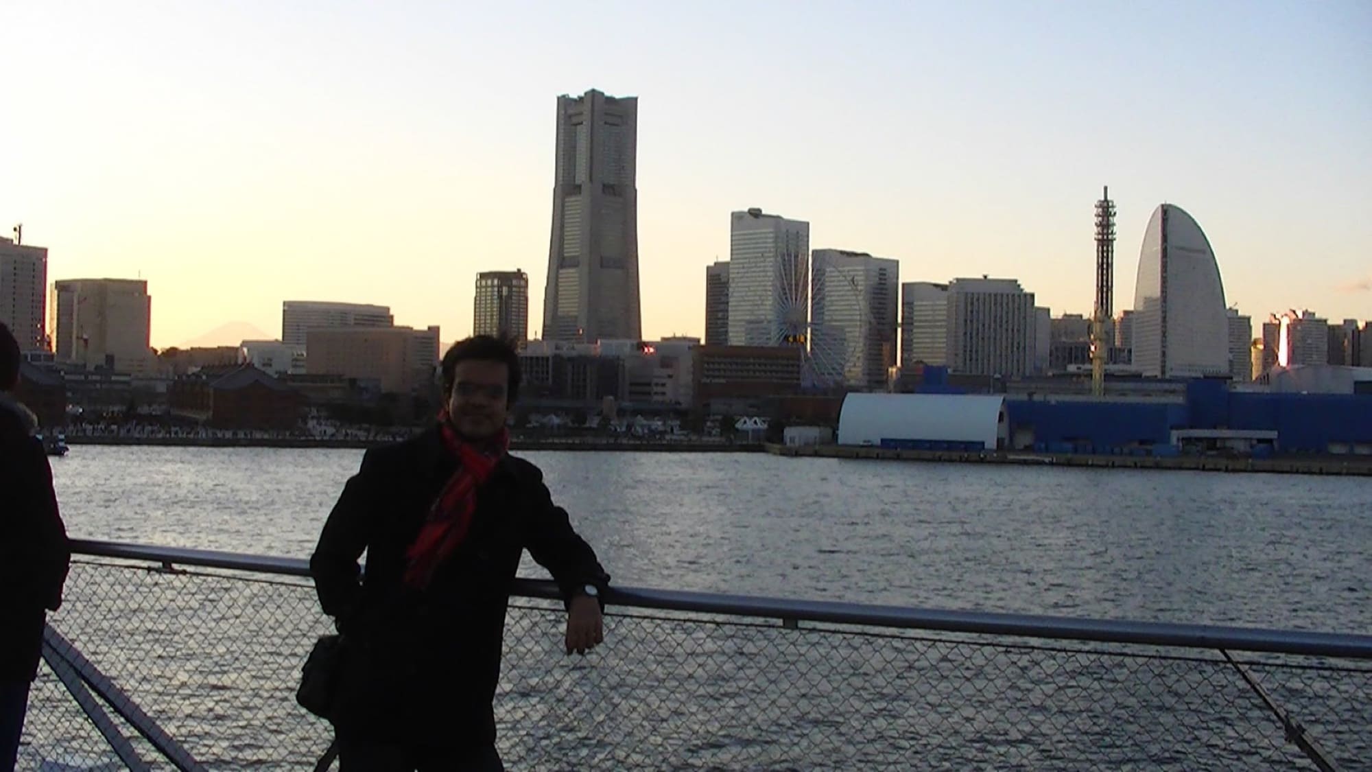 Amit leaning on a harbour railing in Yokohama, the Minato Mirai skyline behind him at sunset.