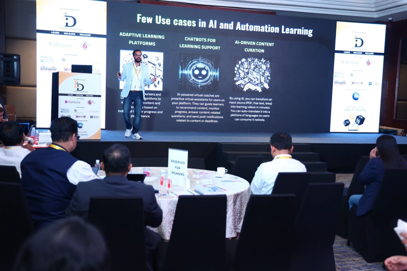 Amit Kumar Soni on a lit stage presenting to a seated audience at round tables.
